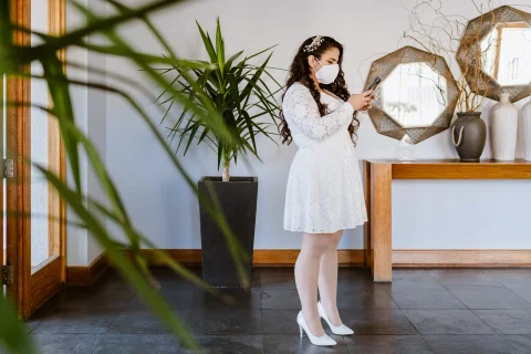 Mujer con vestido blanco y mascarilla mira un teléfono mientras permanece de pie en un vestíbulo.
