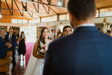 Mujer con vestido blanco y ramo permanece frente a un hombre con traje azul dentro de la iglesia.