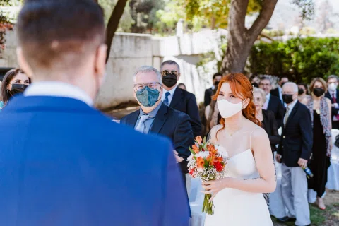 Mujer con vestido blanco y ramo permanece frente a un hombre con traje azul, con otra persona enmascarada detrás.