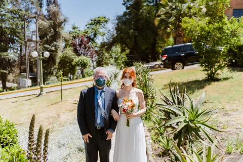 Mujer con vestido blanco sostiene un ramo junto a un hombre con mascarilla en un jardín.