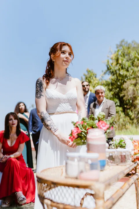 Mujer con vestido blanco permanece junto a una mesa mientras sostiene un ramo.