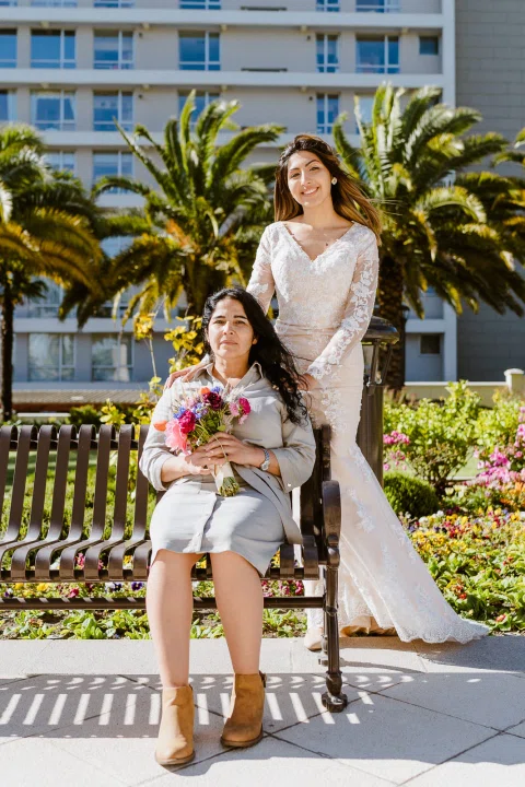 Mujer con vestido blanco permanece de pie detrás de una mujer sentada en una banca que sostiene un ramo de flores.