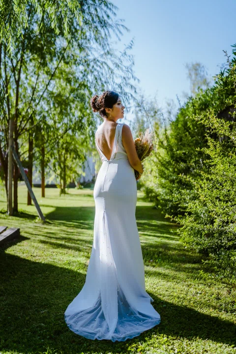Mujer con vestido blanco permanece de pie de espaldas en un sendero del jardín, con el vestido extendido detrás.
