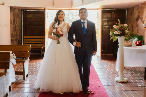 Mujer con vestido blanco entra a la iglesia del brazo de un hombre con traje azul.