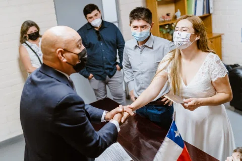 Mujer con vestido blanco estrecha la mano de un hombre con traje mientras sostiene una libreta marrón.