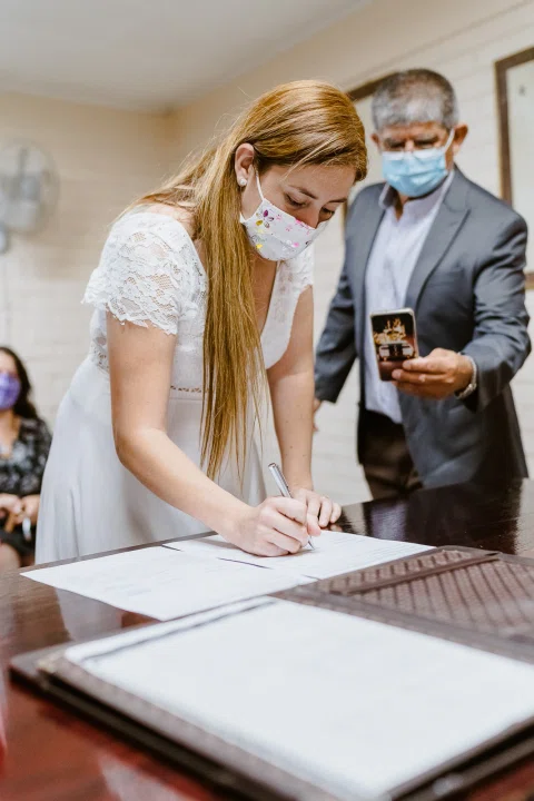 Mujer con vestido blanco inclinada sobre una mesa mientras firma un documento.