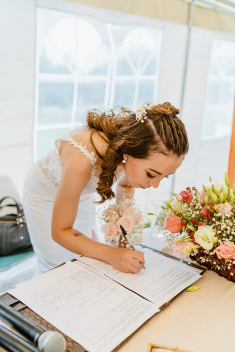 Mujer con vestido blanco inclinada sobre una mesa mientras firma un documento.