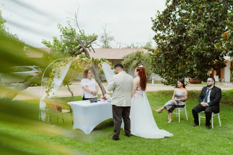 Mujer con vestido blanco permanece frente a un hombre con chaqueta clara y una mujer de cabello rojo junto a una mesa y un arco floral.