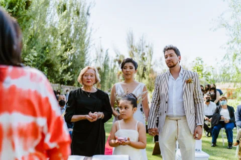 Mujer con vestido blanco y hombre con chaqueta a rayas están junto a una mujer con vestido negro y una niña en un jardín.