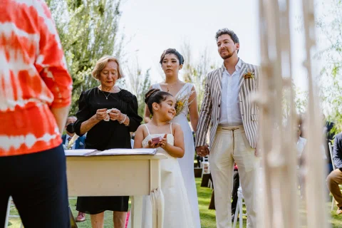 Mujer con vestido blanco, hombre con chaqueta a rayas y una niña están junto a una mesa en un jardín.