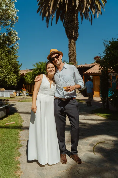Mujer con vestido blanco y hombre con sombrero posan abrazados en un sendero de jardín.