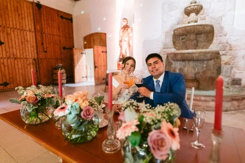 Mujer con vestido blanco y hombre con traje azul están sentados juntos detrás de una mesa decorada con flores.