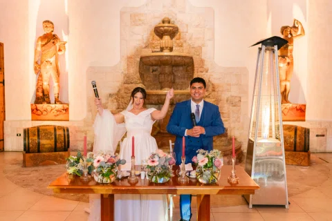 Mujer con vestido blanco y hombre con traje azul posan detrás de una mesa decorada con flores.