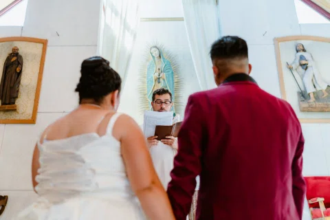 Mujer con vestido blanco y hombre con traje burdeos vistos de espaldas frente al altar.