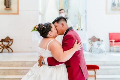 Mujer con vestido blanco y hombre con traje burdeos se besan dentro de una iglesia.