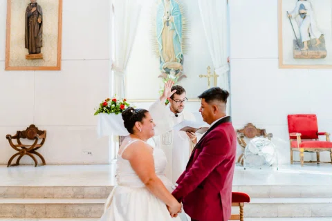 Mujer con vestido blanco y hombre con traje burdeos se miran de frente mientras se toman de la mano.