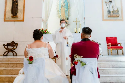 Mujer con vestido blanco y hombre con traje burdeos sentados de espaldas frente al altar.
