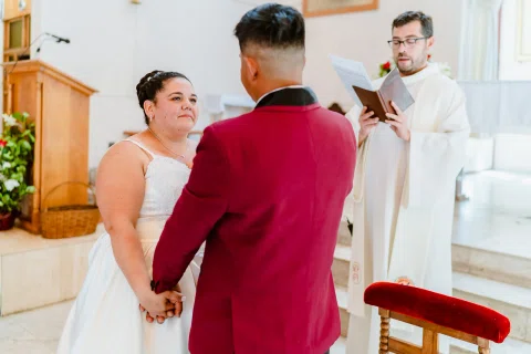 Mujer con vestido blanco y hombre con traje burdeos se toman de la mano frente a una persona con vestimenta blanca que sostiene un libro.