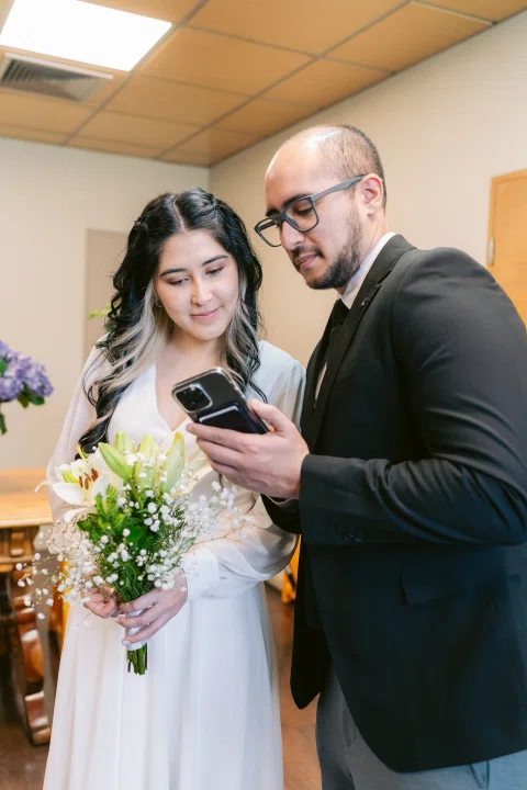 Mujer con vestido blanco y ramo y hombre de traje negro con lentes miran un celular.