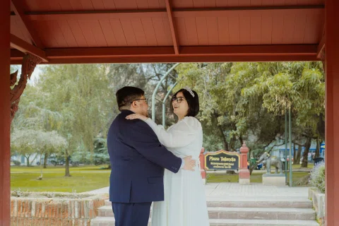 Mujer con vestido blanco y hombre con traje oscuro se abrazan bajo un pabellón de madera.