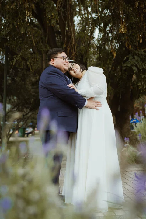 Mujer con vestido blanco y hombre con traje oscuro se besan entre árboles y flores desenfocadas.