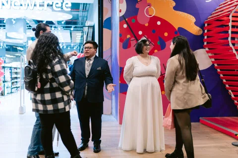 Mujer con vestido blanco y hombre con traje oscuro conversan con dos mujeres frente a un mural colorido.