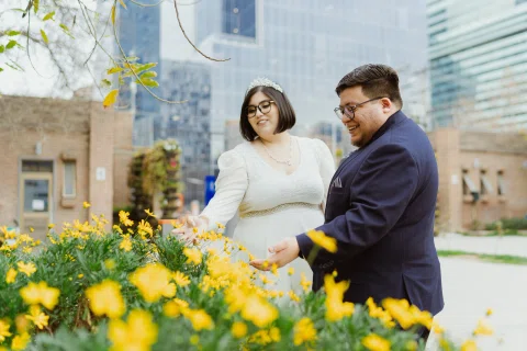 Mujer con vestido blanco y hombre con traje oscuro permanecen junto a flores amarillas, con edificios desenfocados al fondo.