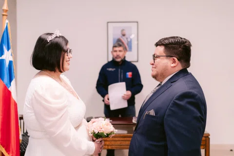 Mujer con vestido blanco y hombre con traje oscuro permanecen frente a frente junto a una bandera chilena; otro hombre sostiene papeles detrás.