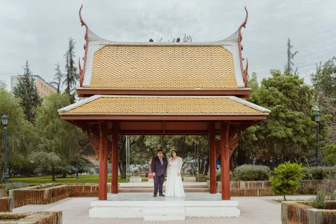 Mujer con vestido blanco y hombre con traje oscuro posan a distancia bajo un pabellón de techo oriental en un parque.