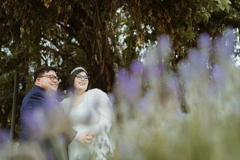 Mujer con vestido blanco y hombre con traje oscuro posan tomados de la mano detrás de flores lilas desenfocadas.