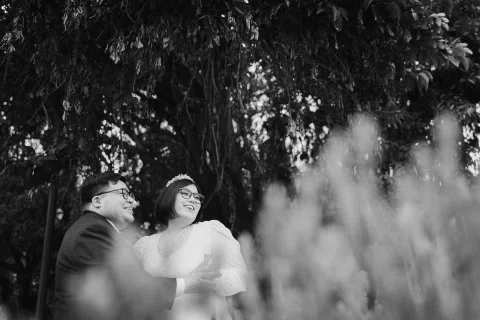 Mujer con vestido blanco y hombre con traje oscuro posan entre vegetación desenfocada, en blanco y negro.
