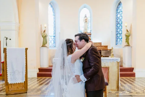 Mujer con vestido blanco y hombre con traje oscuro se besan frente al altar de una iglesia.