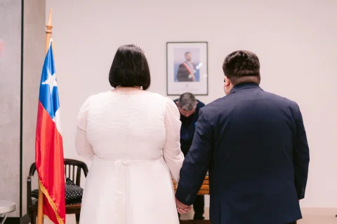 Mujer con vestido blanco y hombre con traje oscuro, vistos de espaldas, permanecen tomados de la mano frente a un hombre detrás de una mesa.