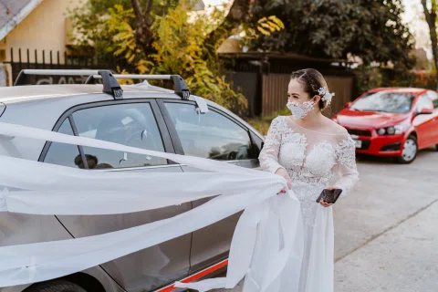 Mujer con vestido blanco y mascarilla permanece junto a un automóvil decorado con una cinta blanca.