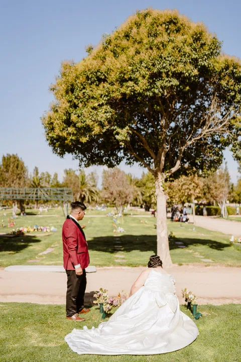 Mujer con vestido blanco está junto a flores y lápidas bajo un árbol mientras un hombre con chaqueta burdeos permanece de pie a corta distancia.