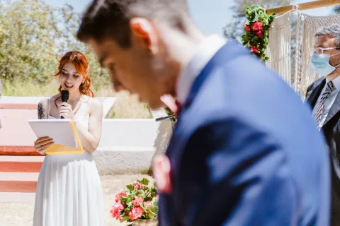 Mujer con vestido blanco lee un documento mientras sostiene un micrófono, con un hombre de traje azul en primer plano.