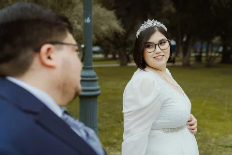 Mujer con vestido blanco, lentes y adorno en el cabello sonríe hacia un hombre con traje oscuro desenfocado en primer plano.