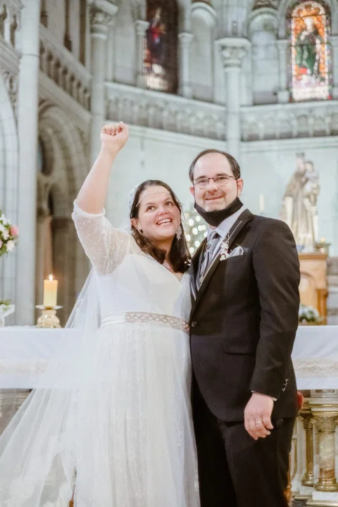 Mujer con vestido blanco sonríe y levanta un brazo junto a un hombre con traje oscuro frente al altar.
