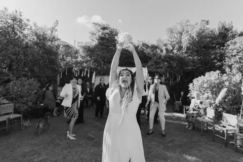 Mujer con vestido blanco levanta un ramo sobre la cabeza frente a un grupo de personas, en blanco y negro.