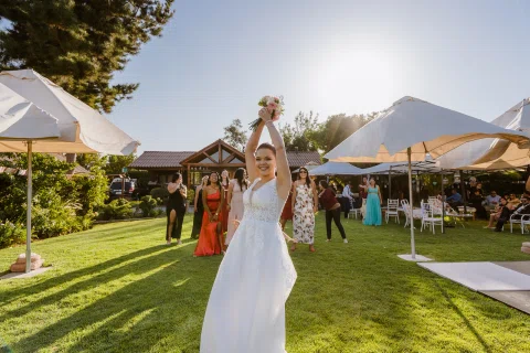 Mujer con vestido blanco levanta un ramo sobre su cabeza en el jardín mientras varias mujeres esperan detrás.
