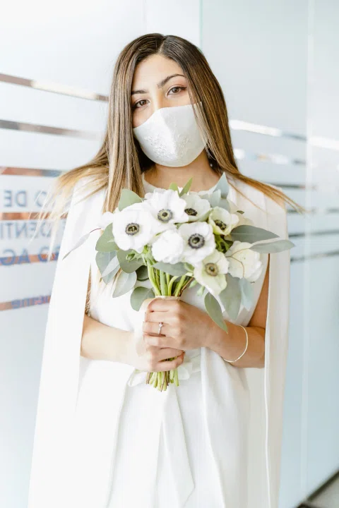 Retrato cercano de una mujer con vestido blanco y mascarilla sosteniendo un ramo de flores blancas.