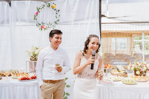 Mujer con vestido blanco sostiene un micrófono y una copa junto a un hombre con camisa blanca que también sostiene una copa, frente a una mesa con comida y flores.