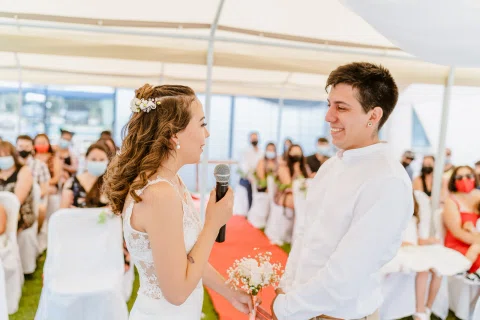 Mujer con vestido blanco sostiene un micrófono frente a un hombre con camisa blanca, con varias personas sentadas detrás.