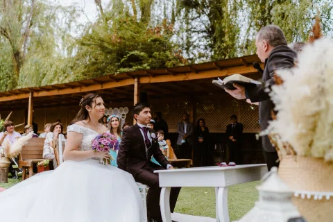 Mujer con vestido blanco mira a un hombre con traje oscuro mientras ambos están sentados junto a una pequeña mesa.
