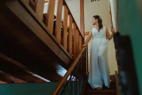 Mujer con vestido blanco permanece sobre una escalera de madera dentro de una casa.