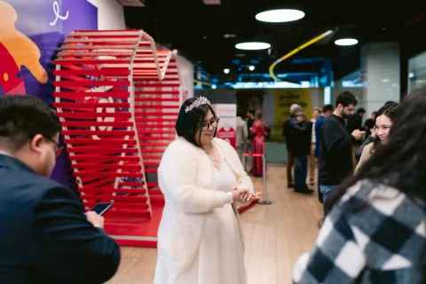 Mujer con vestido blanco permanece junto a una estructura roja mientras mira sus manos en un espacio interior.