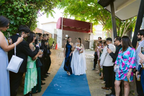 Mujer con vestido blanco y ramo avanza por una alfombra azul entre personas a ambos lados.