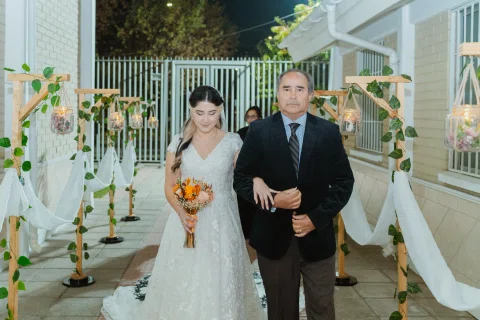Mujer con vestido blanco y ramo naranja camina tomada del brazo de un hombre con traje oscuro.