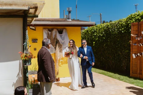 Mujer con vestido blanco y ramo camina junto a un hombre con traje azul frente a una casa amarilla.