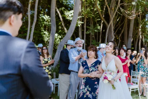 Mujer con vestido blanco y ramo camina junto a una mujer con vestido floral mientras otras personas observan en el jardín.